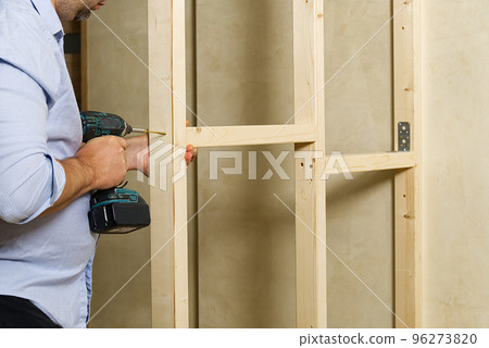 A man fastens two wooden beams with a self-tapping screw. The joiner wraps the screw with a screwdriver. Construction of a timber frame for plasterboard A man fastens two wooden beams with a self-tapping screw. The joiner wraps the screw with a screwdriver. Construction of a timber frame for plasterboard 96273820