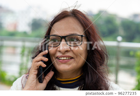 Mid Adult Smiling Woman with long brown hair Speaking on mobile phone outdoors. Closeup. She is wearing White T-shirt and specs. Front view. Head and Shoulder Shot. Garden lake side in the background. Mid Adult Smiling Woman with long brown hair Speaking on mobile phone outdoors. Closeup. She is wearing White T-shirt and specs. Front view. Head and Shoulder Shot. Garden lake side in the background. 96277235