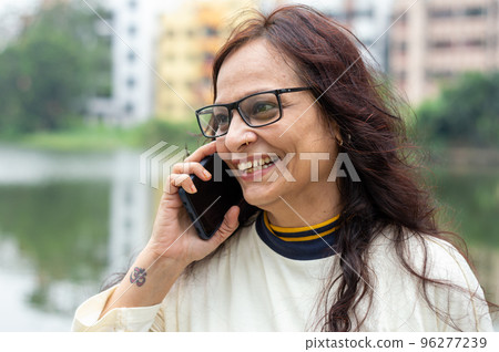 Mid Adult Smiling Woman with long brown hair Speaking on mobile phone outdoors. Closeup. She is wearing White T-shirt and specs. Front view. Head and Shoulder Shot. Garden lake side in the background. Mid Adult Smiling Woman with long brown hair Speaking on mobile phone outdoors. Closeup. She is wearing White T-shirt and specs. Front view. Head and Shoulder Shot. Garden lake side in the background. 96277239