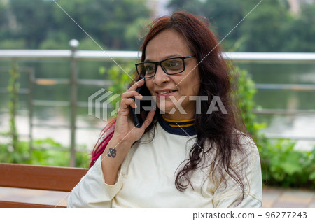 Mid Adult Smiling Woman with long brown hair Speaking on mobile phone outdoors. Closeup. She is wearing White T-shirt and specs. Front view. Head and Shoulder Shot. Garden lake side in the background. 96277243