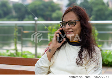 Mid Adult Smiling Woman with long brown hair Speaking on mobile phone outdoors. Closeup. She is wearing White T-shirt and specs. Front view. Head and Shoulder Shot. Garden lake side in the background. Mid Adult Smiling Woman with long brown hair Speaking on mobile phone outdoors. Closeup. She is wearing White T-shirt and specs. Front view. Head and Shoulder Shot. Garden lake side in the background. 96277245