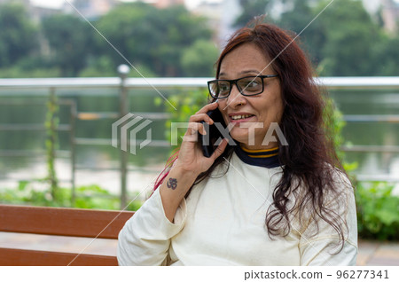 Mid Adult Smiling Woman with long brown hair Speaking on mobile phone outdoors. Closeup. She is wearing White T-shirt and specs. Front view. Head and Shoulder Shot. Garden lake side in the background. 96277341
