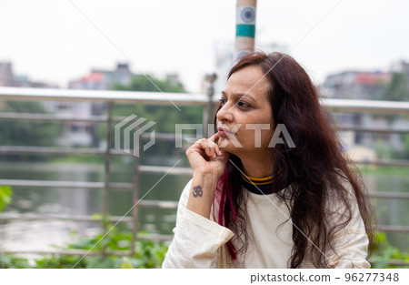 Mid Adult worried Woman sitting on hand on chin pose in a park bench and looking at a distance. Front view. Indian Ethnicity Age 50 to 54 Years. 96277348