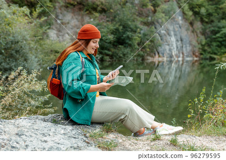 Stylish woman with green backpack holding smart phone and paper map outdoors. Full body photo. Hiking active female lifestyle siting on rocks. Girl wearing red hat relaxing on nature lake background. 96279595