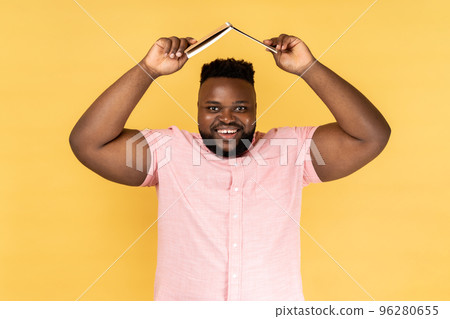Portrait of smiling man wearing pink shirt holding opened book on head, looking at camera with smile, expressing positive emotions. Indoor studio shot isolated on yellow background. Portrait of smiling man wearing pink shirt holding opened book on head, looking at camera with smile, expressing positive emotions. Indoor studio shot isolated on yellow background. 96280655