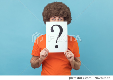 Man with Afro hairstyle in orange T-shirt hiding half of face behind white paper with question mark, finding smart solution, asking for advice. Indoor studio shot isolated on blue background. Man with Afro hairstyle in orange T-shirt hiding half of face behind white paper with question mark, finding smart solution, asking for advice. Indoor studio shot isolated on blue background. 96280656
