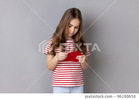 Portrait of concentrated little girl wearing striped T-shirt opening envelope and reading letter with calm expression, got news. Indoor studio shot isolated on gray background. Portrait of concentrated little girl wearing striped T-shirt opening envelope and reading letter with calm expression, got news. Indoor studio shot isolated on gray background. 96280658