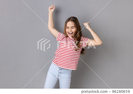 Portrait of joyful little girl wearing striped T-shirt expressing winning gesture with raised fists and screaming, celebrating victory. Indoor studio shot isolated on gray background. Portrait of joyful little girl wearing striped T-shirt expressing winning gesture with raised fists and screaming, celebrating victory. Indoor studio shot isolated on gray background. 96280659