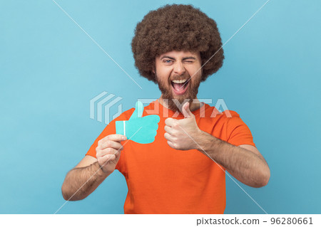 Man with Afro hairstyle wearing orange T-shirt holding and showing like or thumbs up paper shape sign, looking winking at camera with toothy smile. Indoor studio shot isolated on blue background. 96280661