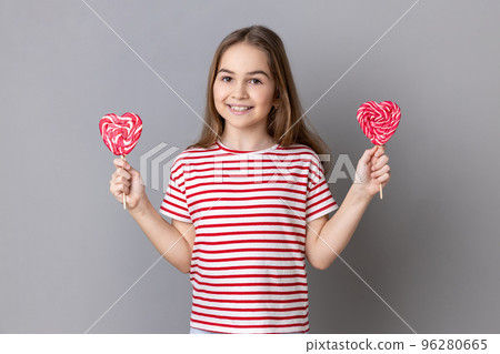 Portrait of happy little girl wearing striped T-shirt tasty heart shape lollypops, having playful expression, looking at camera with smile. Indoor studio shot isolated on gray background. 96280665