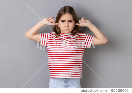 Don't want to listen. Portrait of disobedient little girl wearing striped T-shirt covering ears not to hear advice, ignoring upbringing, parenting. Indoor studio shot isolated on gray background. 96280691