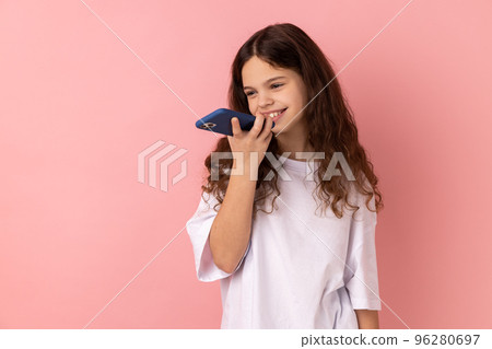 Mobile smart voice technology. Portrait of little girl wearing white T-shirt talking to smartphone using virtual assistant, digital speaker app. Indoor studio shot isolated on pink background. Mobile smart voice technology. Portrait of little girl wearing white T-shirt talking to smartphone using virtual assistant, digital speaker app. Indoor studio shot isolated on pink background. 96280697