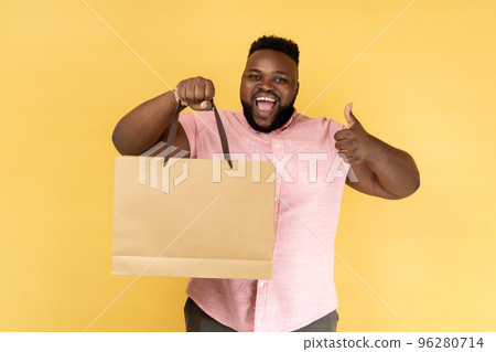 Portrait of man wearing pink shirt showing thumb up holding shopping bag, pleased with good shopping and discounts, recommending. Indoor studio shot isolated on yellow background. 96280714