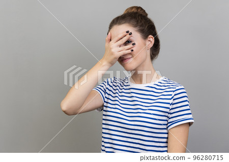Portrait of interested woman wearing striped T-shirt covering eyes with hand, peeking through fingers with happy curious expression. Indoor studio shot isolated on gray background. Portrait of interested woman wearing striped T-shirt covering eyes with hand, peeking through fingers with happy curious expression. Indoor studio shot isolated on gray background. 96280715