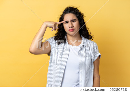 Portrait of woman with dark wavy hair making stupid sign with finger near head, gesturing bad mind, dumb insane idea, frowning face. Indoor studio shot isolated on yellow background. 96280718