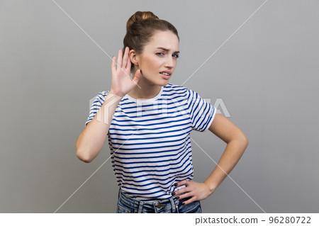 Portrait of nosy woman wearing striped T-shirt listening attentively holding hand near ear to hear confidential talk, hard to understand. Indoor studio shot isolated on gray background. Portrait of nosy woman wearing striped T-shirt listening attentively holding hand near ear to hear confidential talk, hard to understand. Indoor studio shot isolated on gray background. 96280722
