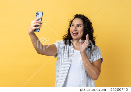 Portrait of friendly woman with dark wavy hair holding smart phone making selfie or making video call, waving hello gesture with hand. Indoor studio shot isolated on yellow background. 96280740