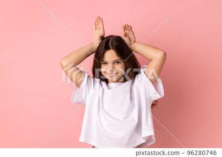 Portrait of positive little girl wearing white T-shirt making funny bunny ears with hands on head, childish behavior, playful optimistic mood. Indoor studio shot isolated on pink background. Portrait of positive little girl wearing white T-shirt making funny bunny ears with hands on head, childish behavior, playful optimistic mood. Indoor studio shot isolated on pink background. 96280746