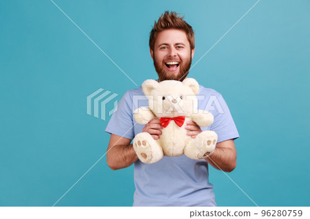 Portrait of happy optimistic handsome bearded man standing looking at camera holding white soft teddy bear, expressing positive emotions. Indoor studio shot isolated on blue background. 96280759