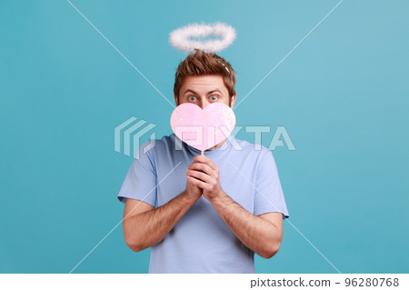 Portrait of angelic man with holy nimbus covering half of face with pink paper heart on stick, being shy to demonstrate love care, romantic feelings. Indoor studio shot isolated on blue background. Portrait of angelic man with holy nimbus covering half of face with pink paper heart on stick, being shy to demonstrate love care, romantic feelings. Indoor studio shot isolated on blue background. 96280768