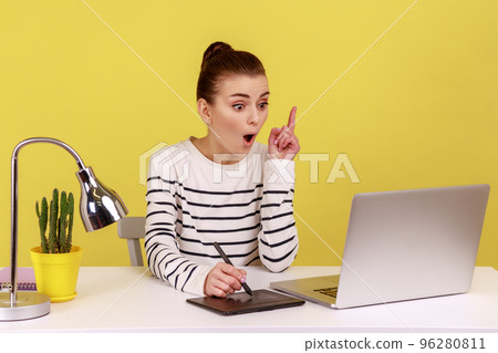 Excited woman designer pointing finger up, having idea, drawing with stylus on graphic tablet, sitting in office working on new collection. Indoor studio studio shot isolated on yellow background. Excited woman designer pointing finger up, having idea, drawing with stylus on graphic tablet, sitting in office working on new collection. Indoor studio studio shot isolated on yellow background. 96280811