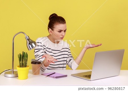 Woman manager sitting at workplace with laptop and looking at laptop screen with angry indignant expression, having conflict with coworkers. Indoor studio studio shot isolated on yellow background. Woman manager sitting at workplace with laptop and looking at laptop screen with angry indignant expression, having conflict with coworkers. Indoor studio studio shot isolated on yellow background. 96280817
