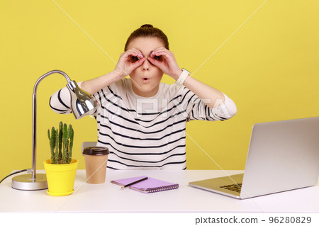 Astonished shocked woman office worker sitting at workplace holding hands near eyes pretending to look in binoculars, find something surprised. Indoor studio studio shot isolated on yellow background. Astonished shocked woman office worker sitting at workplace holding hands near eyes pretending to look in binoculars, find something surprised. Indoor studio studio shot isolated on yellow background. 96280829