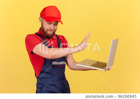 Side view portrait of handsome worker man wearing blue uniform and red cap holding notebook, forbidden information, showing stop ban gesture. Indoor studio shot isolated on yellow background. Side view portrait of handsome worker man wearing blue uniform and red cap holding notebook, forbidden information, showing stop ban gesture. Indoor studio shot isolated on yellow background. 96280845