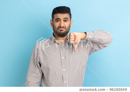 Portrait of young adult businessman criticizing bad quality with thumbs down displeased grimace, showing dislike gesture, wearing striped shirt. Indoor studio shot isolated on blue background. 96280849