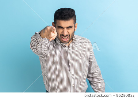 Serious bearded businessman pointing finger, indicating direction to camera and looking at camera with anger, making choice, wearing striped shirt. Indoor studio shot isolated on blue background. Serious bearded businessman pointing finger, indicating direction to camera and looking at camera with anger, making choice, wearing striped shirt. Indoor studio shot isolated on blue background. 96280852