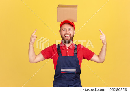 Portrait of excited courier man wearing blue uniform standing pointing at cardboard box on his head, looking at camera with happy expression. Indoor studio shot isolated on yellow background. 96280858