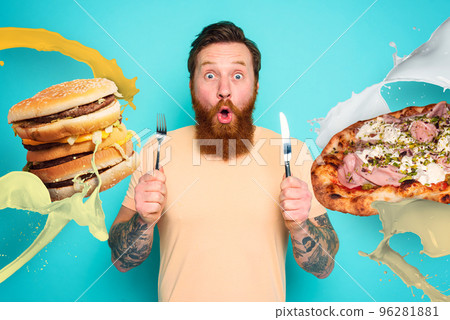 Man with tattoos is ready to eat sandwich and pizza with cutlery in hand. cyan background Man with tattoos is ready to eat sandwich and pizza with cutlery in hand. cyan background 96281881