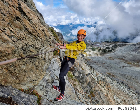 Via Ferrata climbing guided tour excursion happy Asian woman tourist hanging from rope at high pass on mountain side on Whistler peak, BC, Canada Travel 96282041
