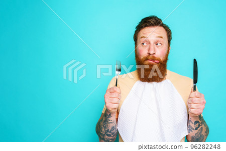 Man with tattoos is ready to eat with cutlery in hand. cyan background 96282248