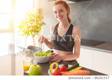 Athletic young red haired woman in the home kitchen eating a healthy salad 96282317