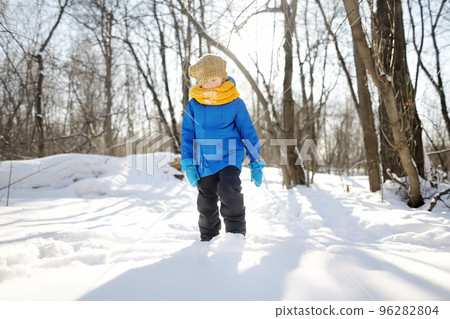 Little boy having fun in forest on sunny winter day. Child dressed in a warm clothes, hat, hand gloves and scarf. Active outdoors leisure for kids on nature among driven snow 96282804