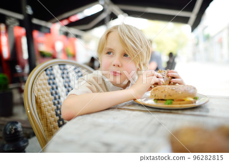 Cute blonde boy eating large bagel with salmon and arugula in outdoor fast food restaurant Cute blonde boy eating large bagel with salmon and arugula in outdoor fast food restaurant 96282815