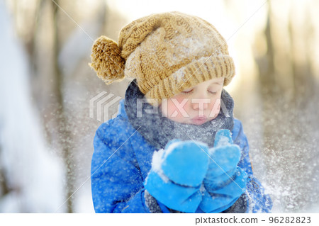 Little boy blowing snow from his hands. Child enjoy walking in the park on snowy day. Baby having fun during snowfall. Outdoor winter activities for kids. 96282823