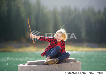 Cute school child is playing with stick and having fun on the coast of mountain lake. Little boy travel in the National Park Durmitor, Montenegro. Summer vacation activity for kids. 96282901
