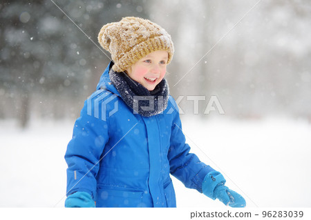 Little boy having fun playing with fresh snow during snowfall. Baby catching snowflakes on gloves. Kid dressed in warm clothes, hat, hand gloves and scarf. Active winter outdoors leisure for child 96283039