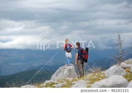 Cute schoolchild and his mature father hiking together on mountain and exploring nature. Child watching a clouds. Concepts of adventure, scouting and hiking tourism for kids. 96283181
