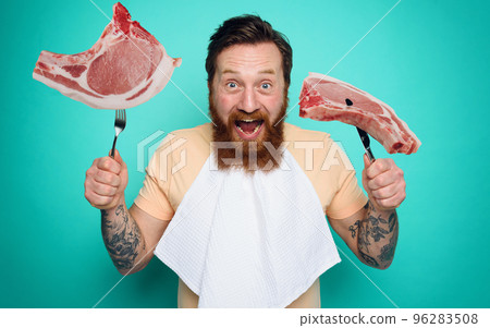 Man with tattoos is ready to eat meat with cutlery in hand. cyan background 96283508