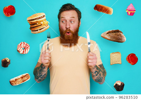 Man with tattoos is ready to eat sandwich and sweets with cutlery in hand. cyan background 96283510