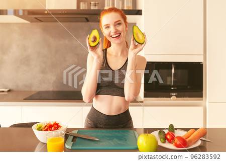 Young red hair woman smiles showing an avocado while preparing a snack in the kitchen Young red hair woman smiles showing an avocado while preparing a snack in the kitchen 96283592