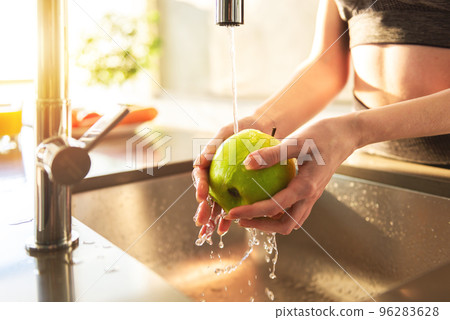 Young woman washes, with running water, an apple in the kitchen sink illuminated by the sun 96283628