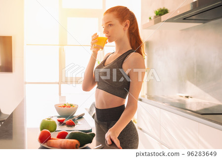 Athletic young red haired woman in the kitchen drinks a glass of fruit centrifuged juice Athletic young red haired woman in the kitchen drinks a glass of fruit centrifuged juice 96283649