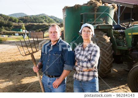 Senior spouses farmers posing near farming equipment 96284143