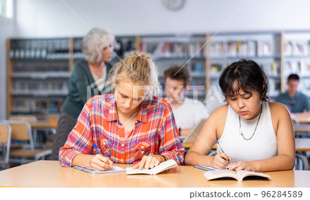Two teenage friends sit at a desk in school library and carefully do assignments Two teenage friends sit at a desk in school library and carefully do assignments 96284589