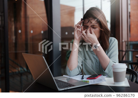 A tired and stressed young woman holds glasses and massages his nose while working hard in a cafe. A tired and stressed young woman holds glasses and massages his nose while working hard in a cafe. 96286170