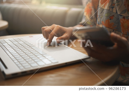 Close-up of woman's hand using smartphone while working on laptop in a coffee shop. Close-up of woman's hand using smartphone while working on laptop in a coffee shop. 96286173